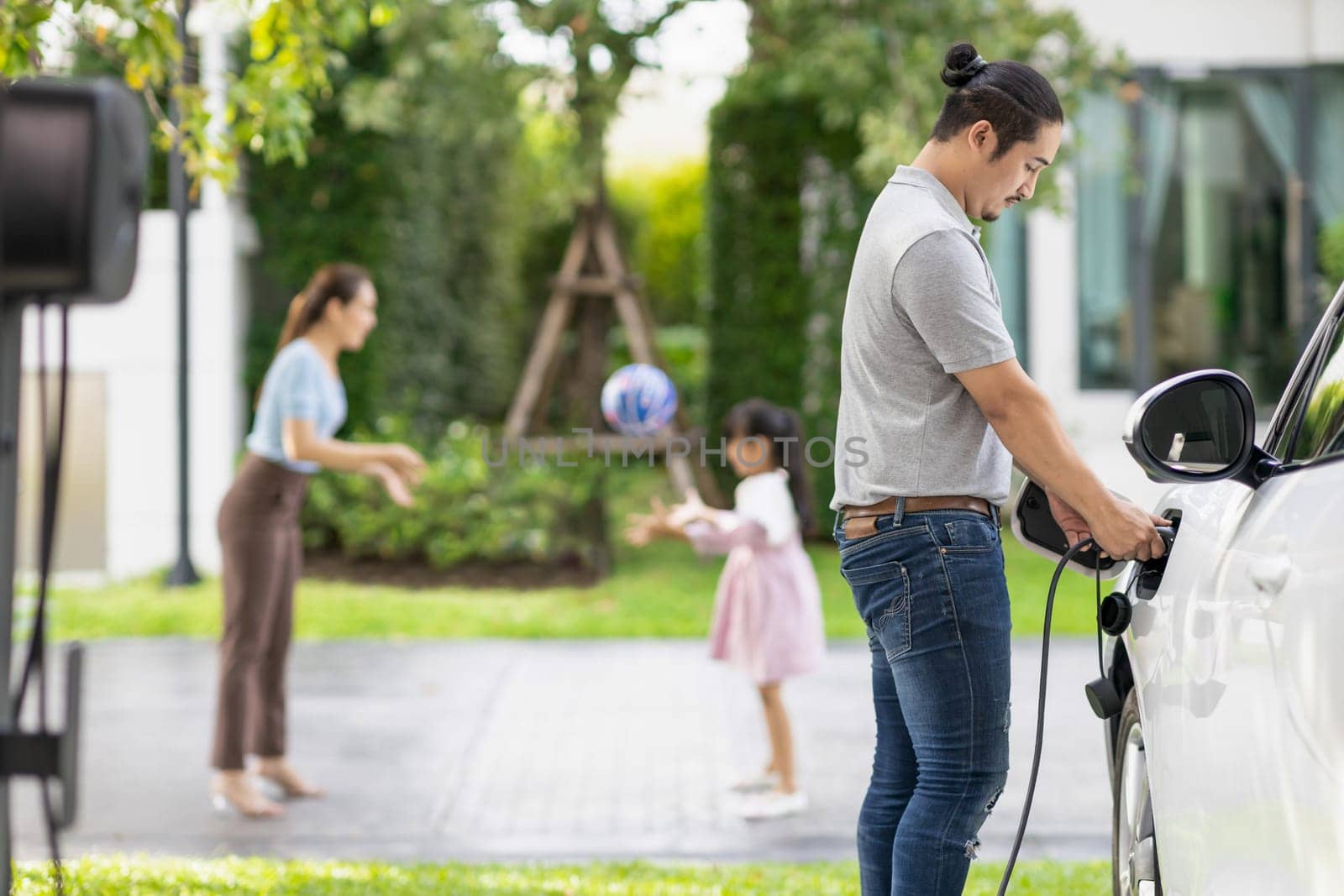 Focus image of progressive man charging electric car from home charging station with blur mother and daughter playing together in the background.