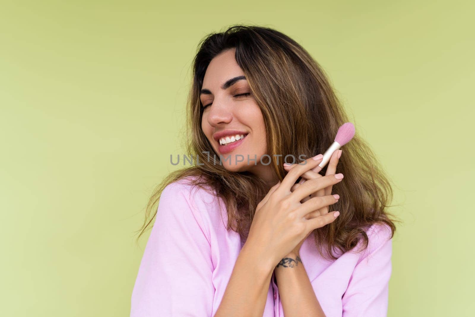 Young woman in casual wear isolated on green background playful positive with blush brush, natural daily makeup