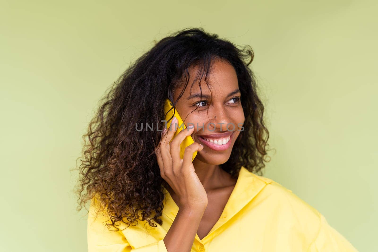 Beautiful african american woman in casual shirt on green background talk on mobile phone smile and laugh