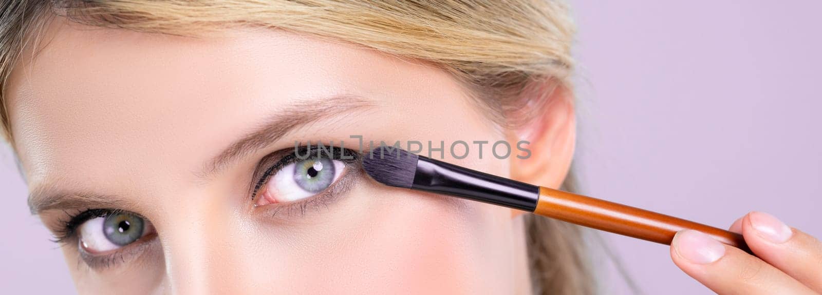 Closeup beautiful girl with flawless applying alluring eye shadow makeup with eyeliner brush. Cosmetic facial painting process on lovely young woman with perfect clean skin on pink isolated background