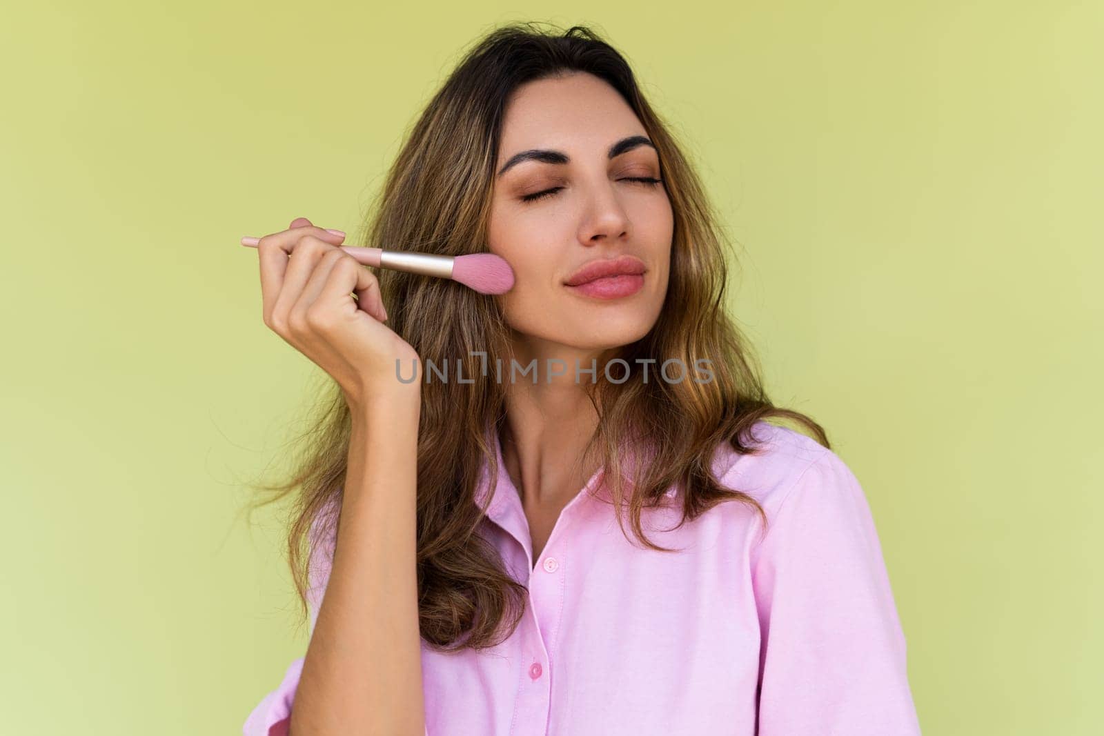 Young woman in casual wear isolated on green background playful positive with blush brush, natural daily makeup by kroshka_nastya