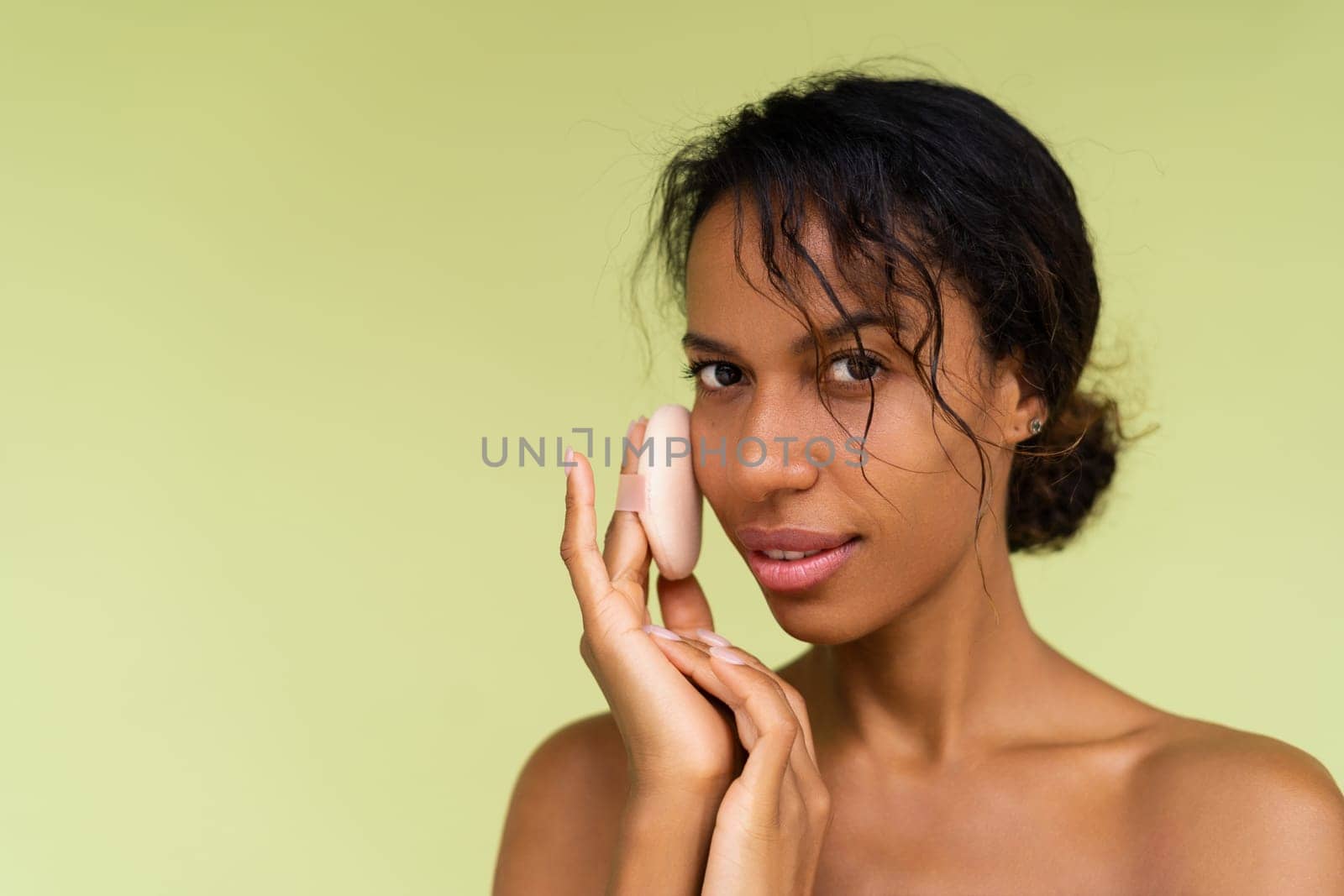 Beauty portrait of young topless african american woman with bare shoulders on green background with cosmetic powder puff velour makeup foundation blender sponge applicator