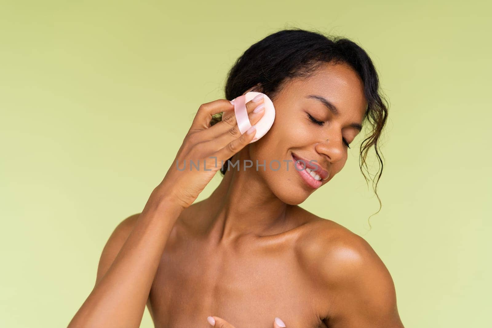 Beauty portrait of young topless african american woman with bare shoulders on green background with cosmetic powder puff velour makeup foundation blender sponge applicator