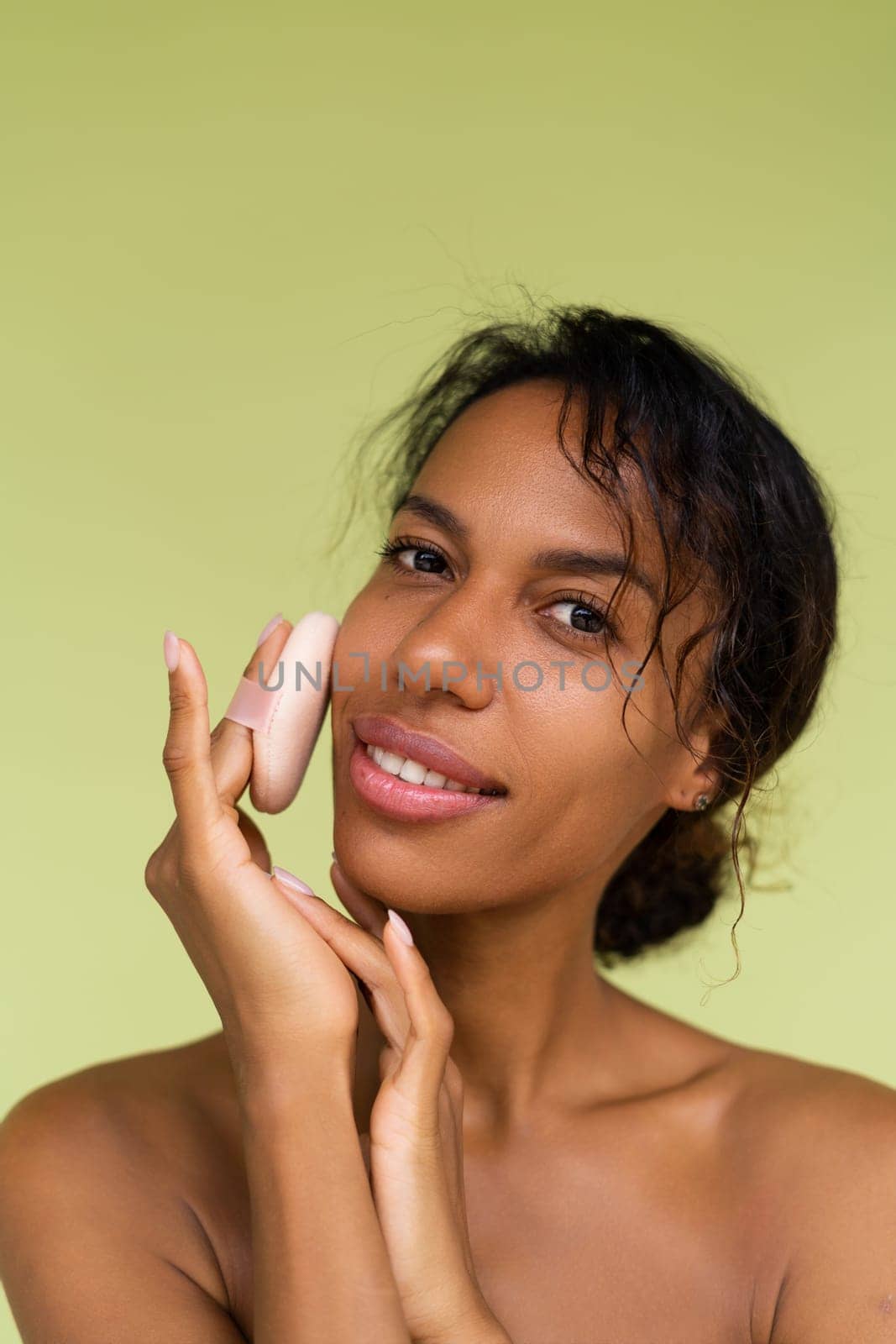 Beauty portrait of young topless african american woman with bare shoulders on green background with cosmetic powder puff velour makeup foundation blender sponge applicator