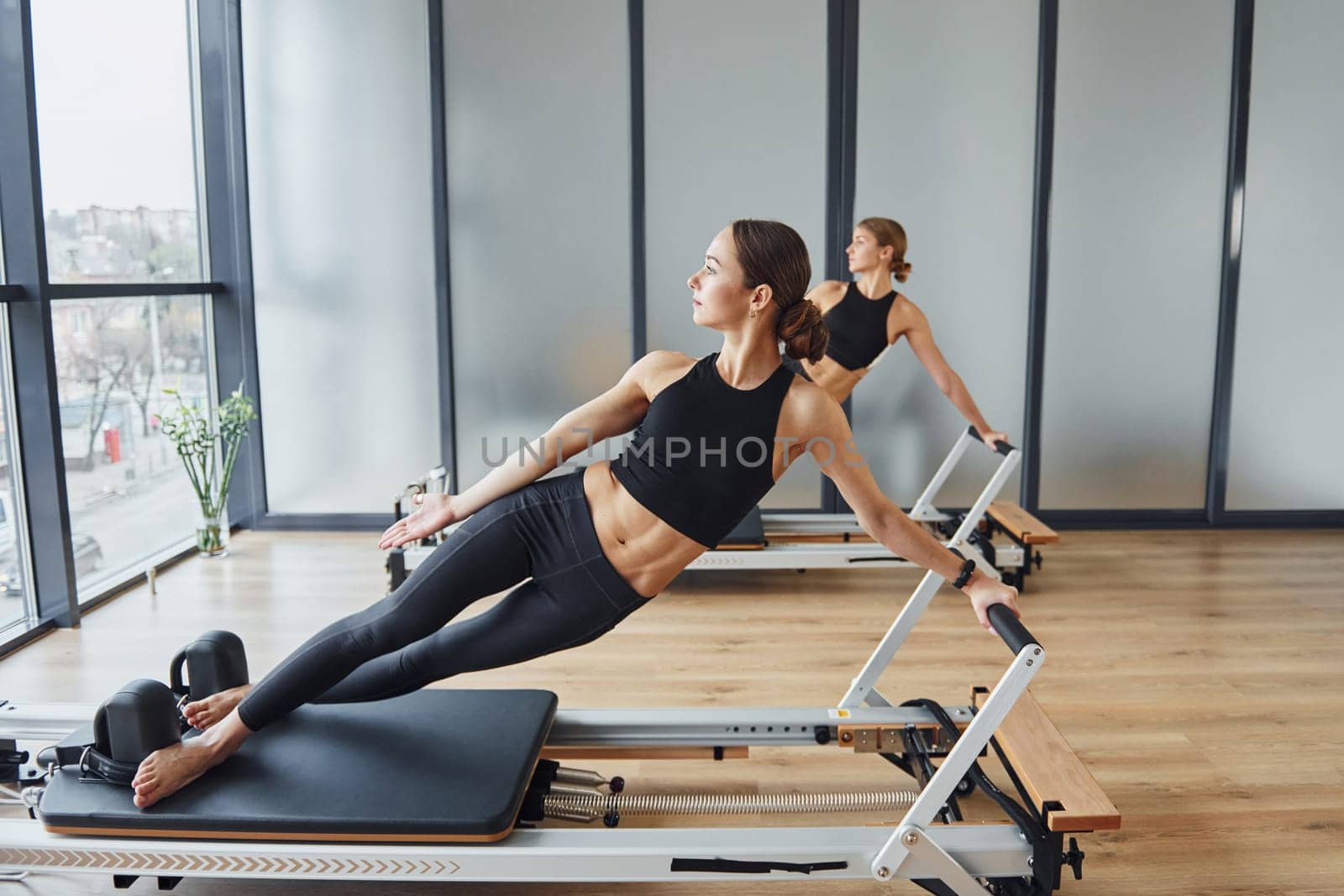 Standing on gym equipment and doing stretches. Two women in sportive wear and with slim bodies have fitness yoga day indoors together.