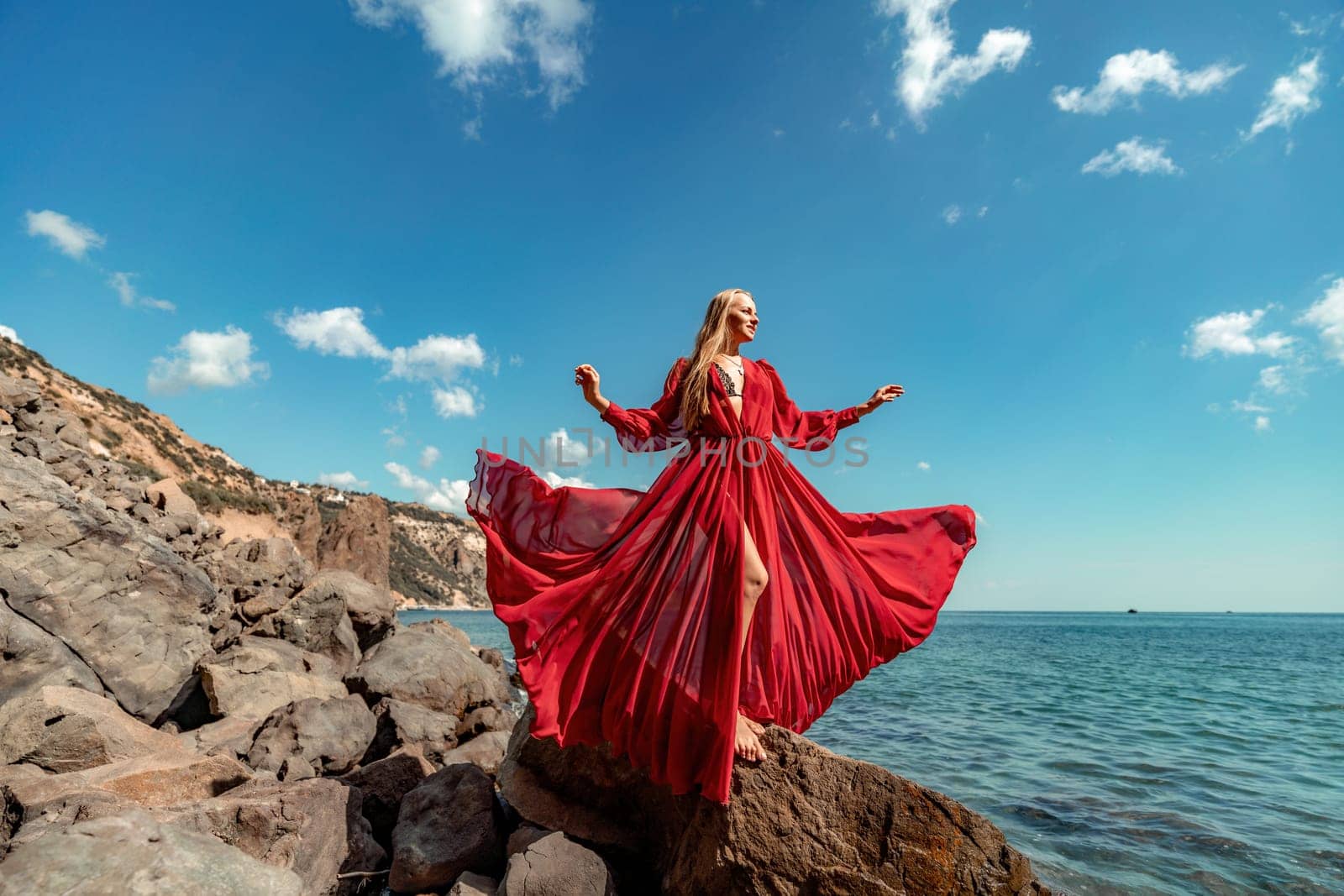 Red dress sea woman. A blonde with flowing hair in a long flowing red dress stands on a rock near the sea. Travel concept, photo session at sea by Matiunina