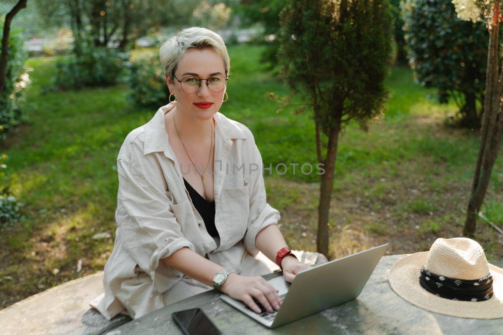 woman freelancer in glasses works at a computer at a white table in nature and spends her day productively by Matiunina