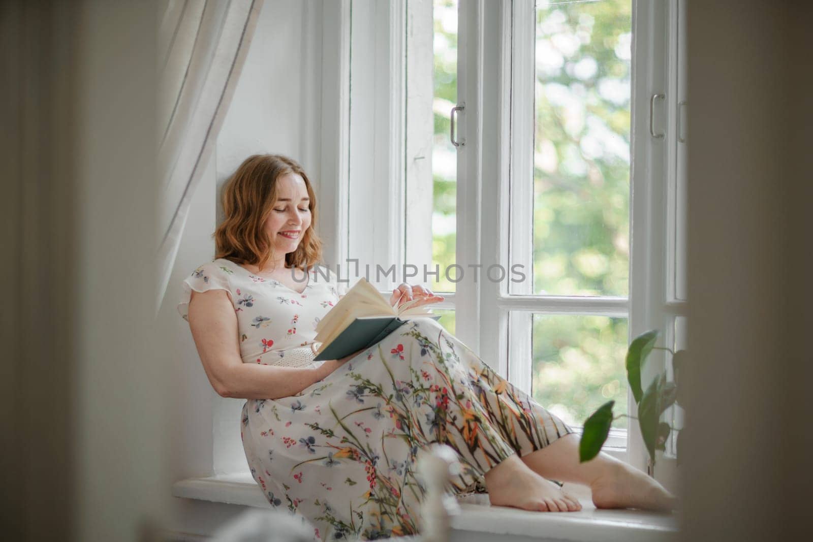 A middle-aged woman in a cream dress sits mysteriously and looks out the window on the windowsill. Green trees outside