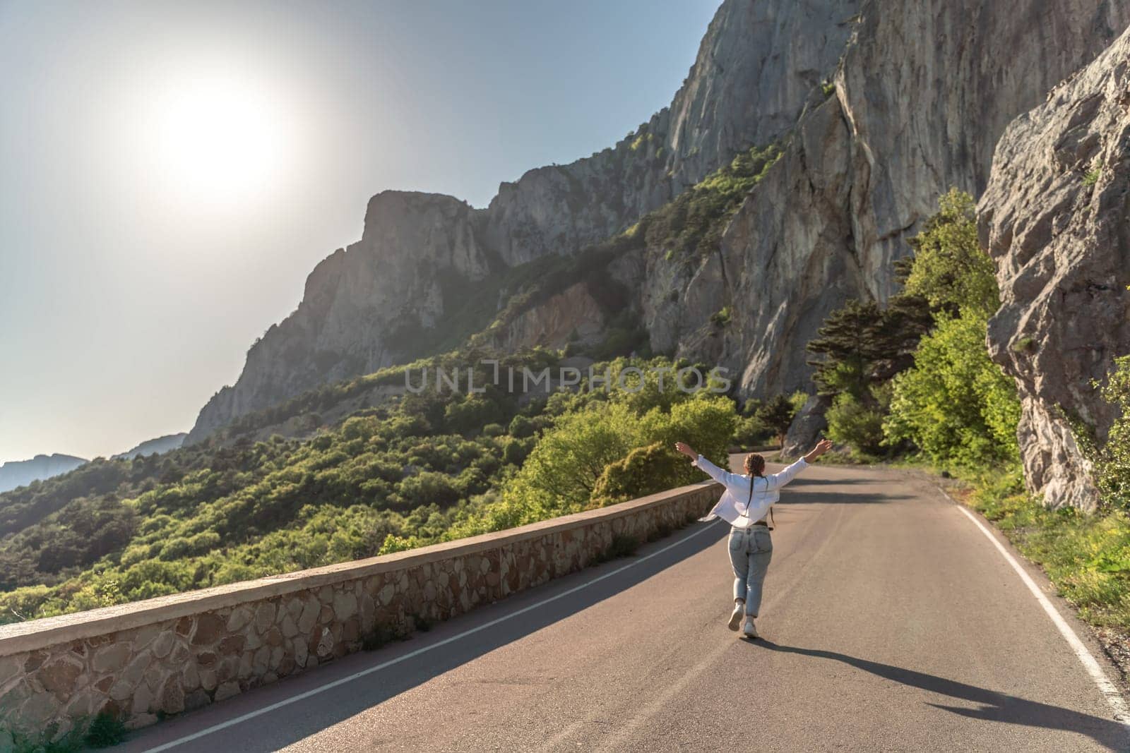 A woman runs along an asphalt road in the middle of a mountainous area. She is dressed in jeans and a white shirt, her hair is braided. A traveler in the mountains rejoices in nature