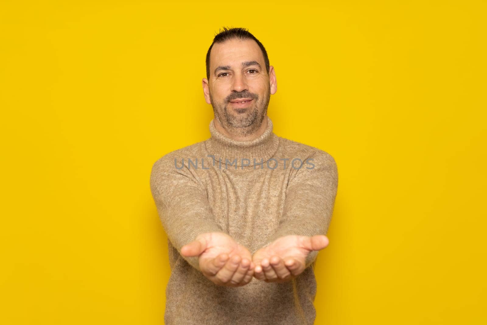 Bearded hispanic handsome man dressed in a beige turtleneck offering something in his hands, has a friendly and receptive attitude. Isolated on yellow background.