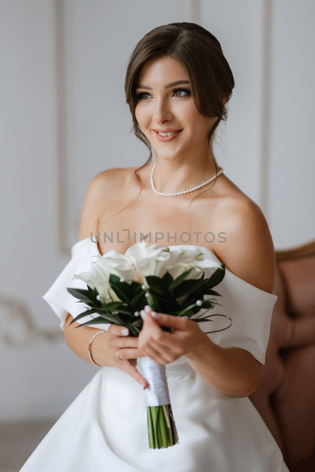 brunette bride in a tight wedding dress in a bright studio with a bouquet on an armchair