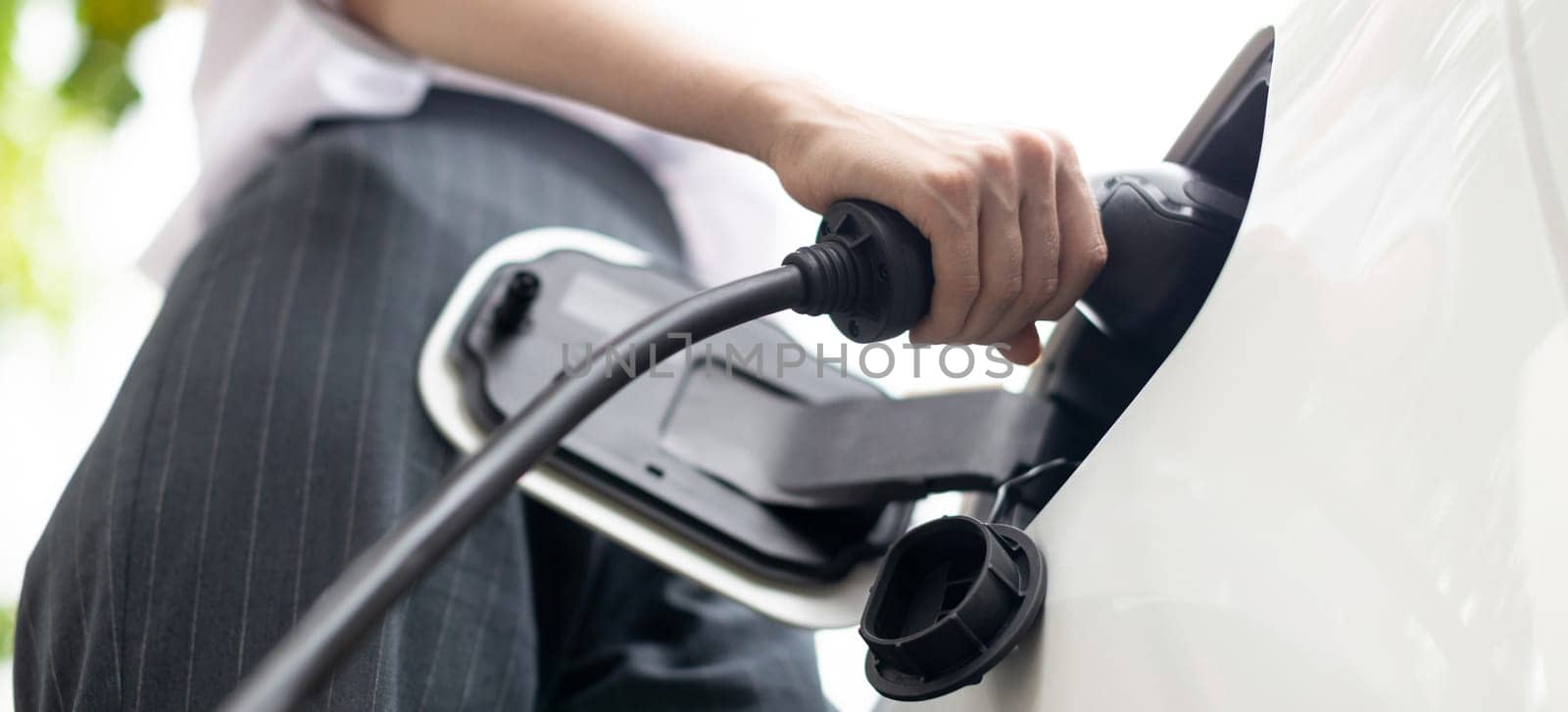 Focus EV charger plug and electric car at public charging station with blur progressive businesswoman holding charger and apartment condo building in background. Eco friendly electric vehicle concept.