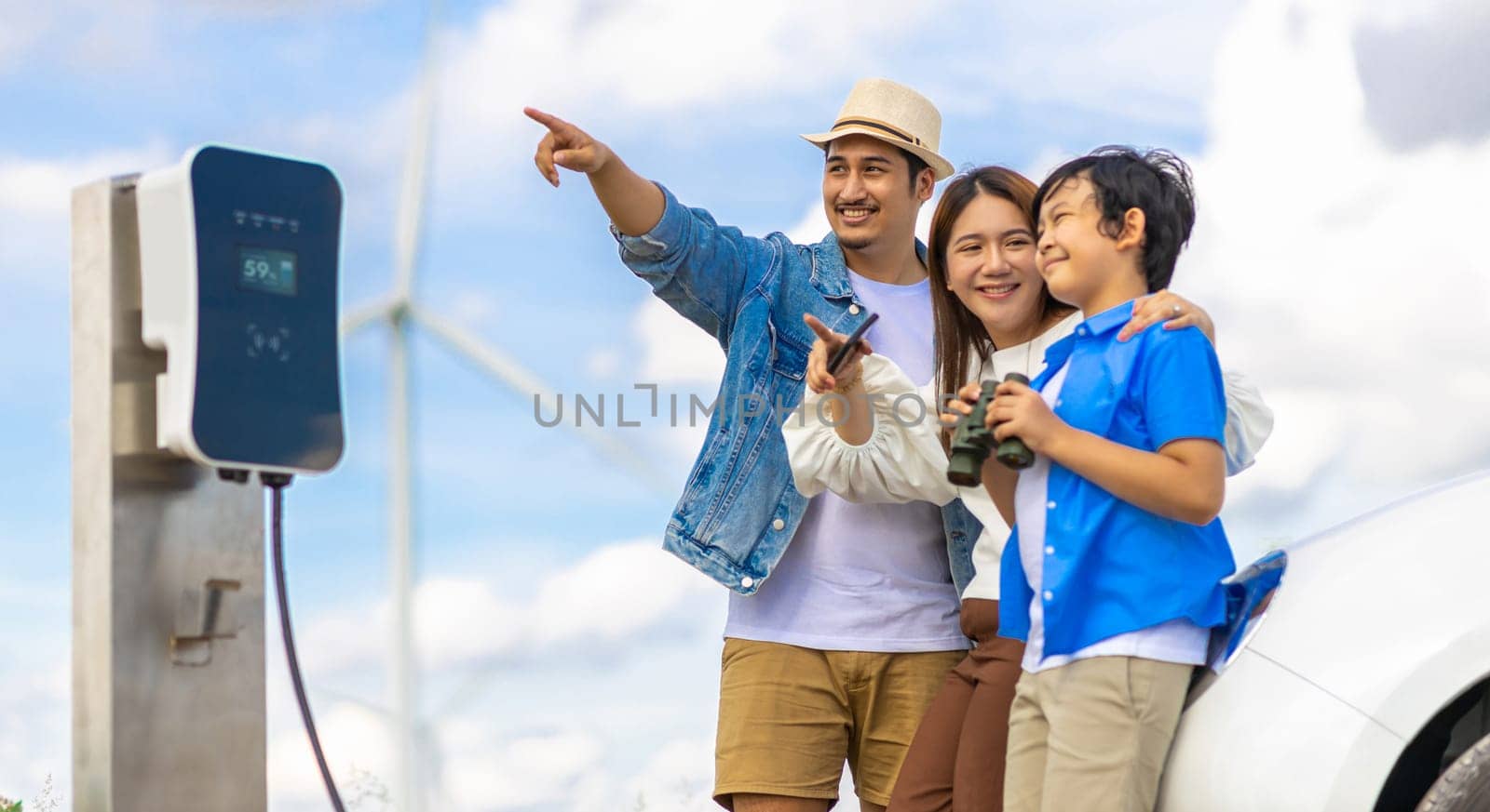 Concept of progressive happy family enjoying their time at wind farm with electric vehicle. Electric vehicle driven by clean renewable energy from wind turbine generator for charging station.