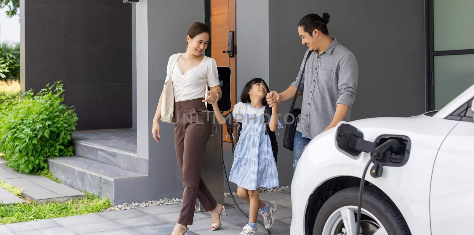 Progressive young parents and daughter with electric vehicle and home charging station. Green and clean energy from electric vehicles for healthy environment. Eco power from renewable source at home.