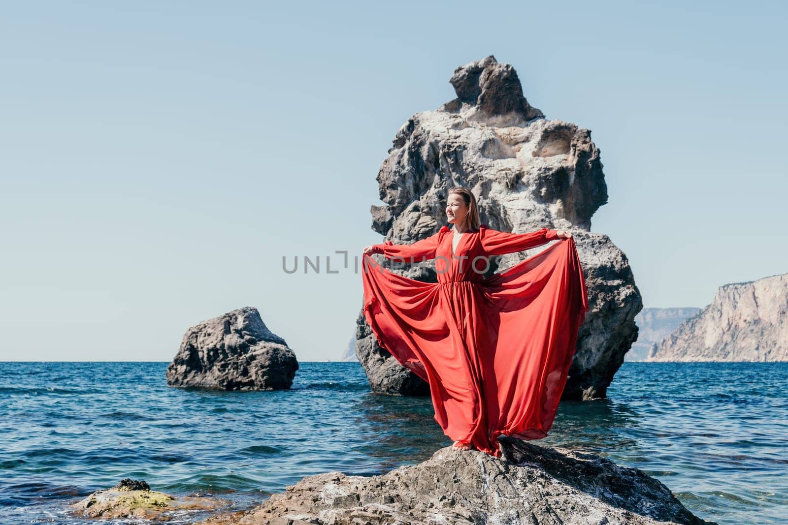 Woman travel sea. Young Happy woman in a long red dress posing on a beach near the sea on background of volcanic rocks, like in Iceland, sharing travel adventure journey