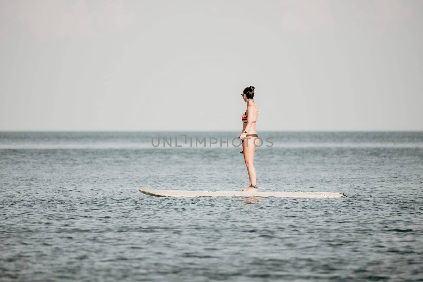 Sea woman sup. Silhouette of happy middle aged woman in rainbow bikini, surfing on SUP board, confident paddling through water surface. Idyllic sunset. Active lifestyle at sea or river