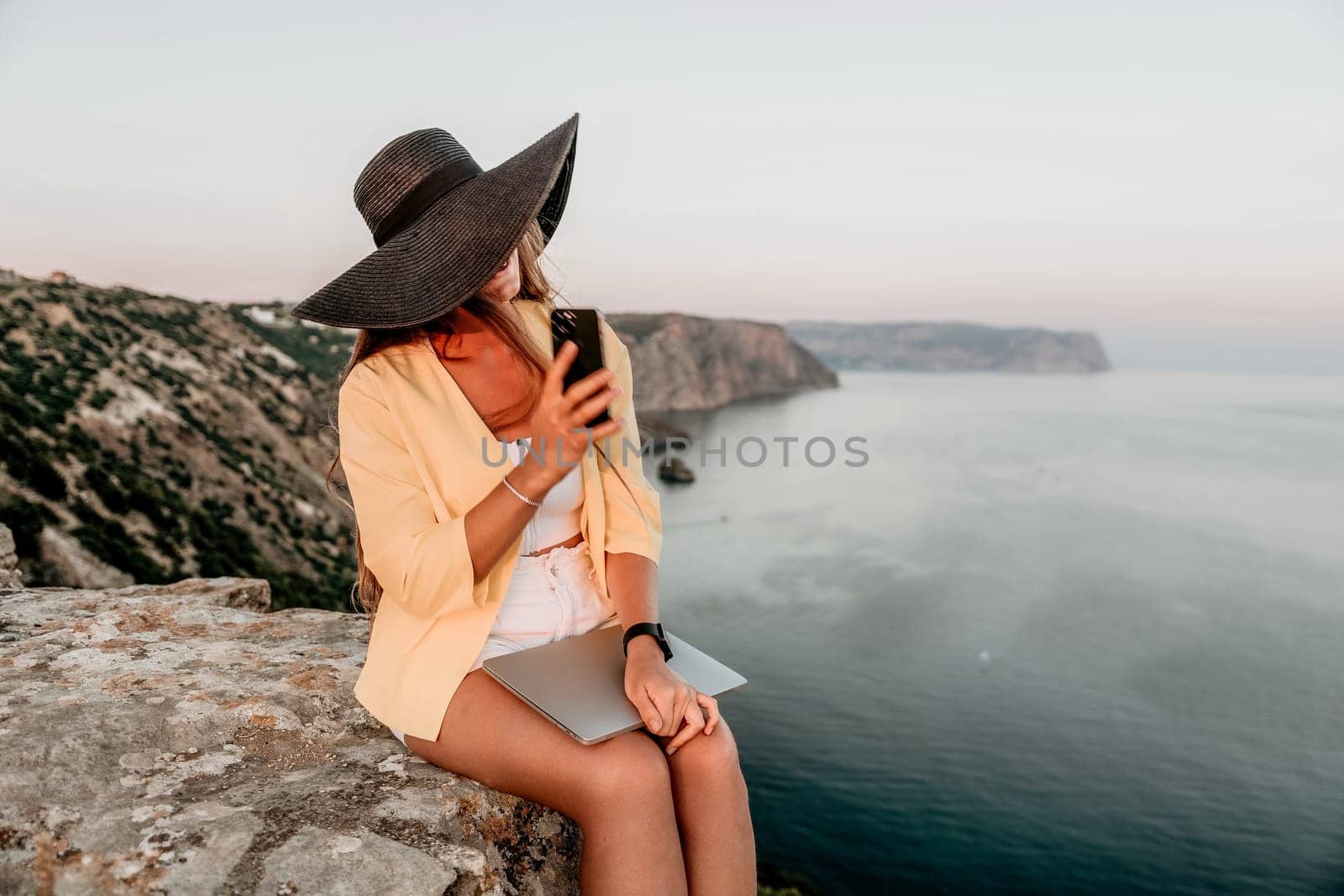 Successful business woman in yellow hat working on laptop by the sea. Pretty lady typing on computer at summer day outdoors. Freelance, travel and holidays concept.