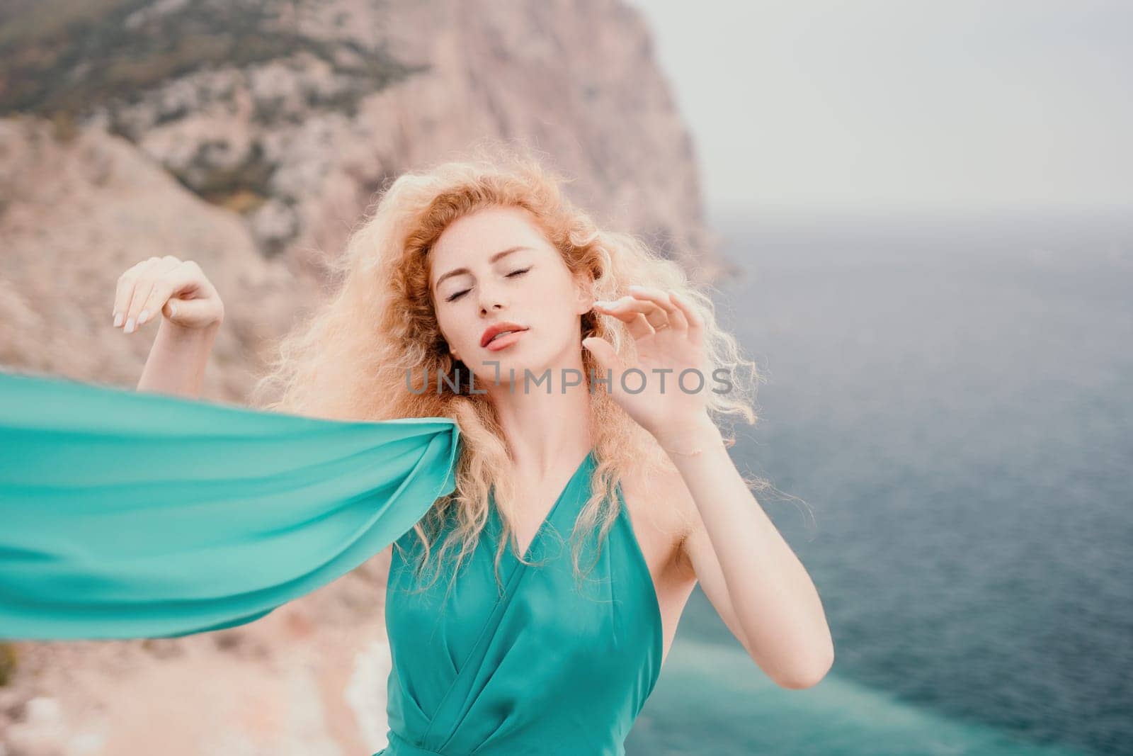 Side view a Young beautiful sensual woman in a mint long dress posing on a volcanic rock high above the sea during sunset. Girl on the nature on overcast sky background. Fashion photo