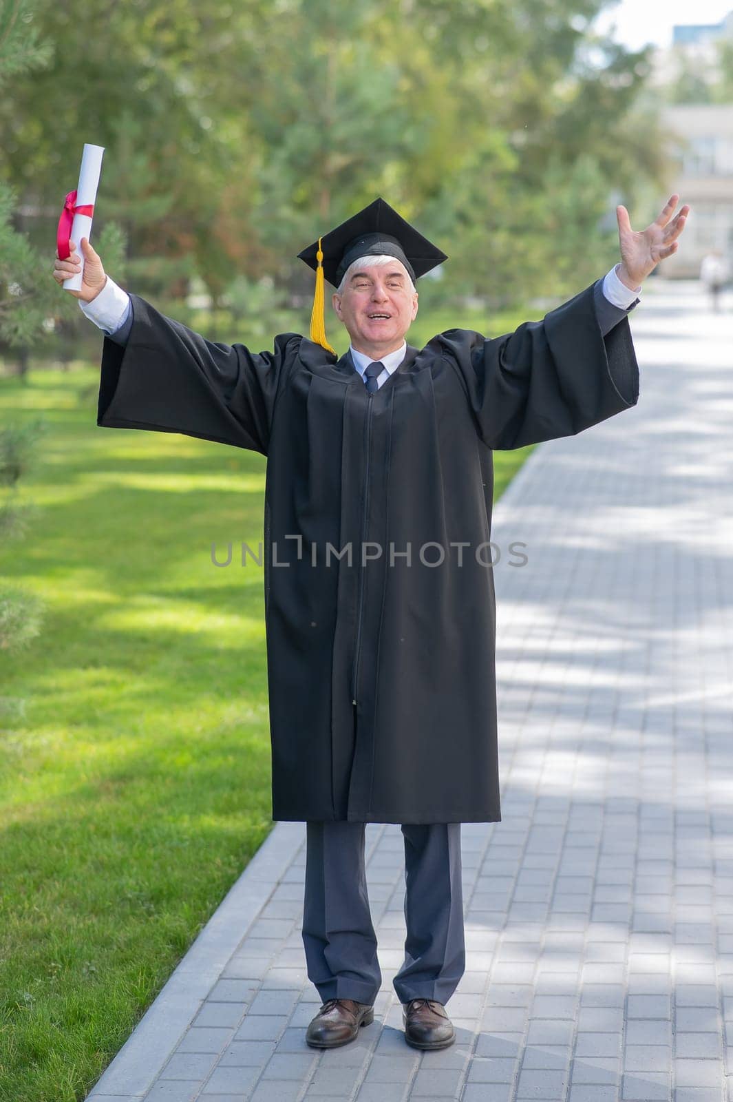 An elderly gray-haired man in a graduate robe spread his arms to the sides and holds a diploma outdoors. Vertical. by mrwed54