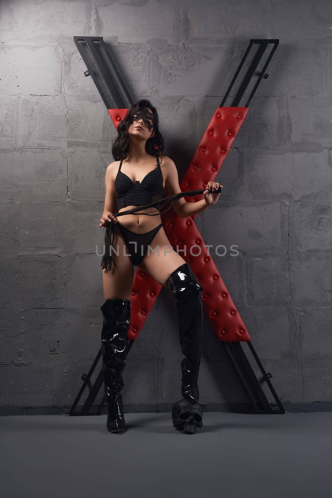 Full body of sexy dominatrix wearing black latex boots with mask standing on skull with whip in hands near X cross