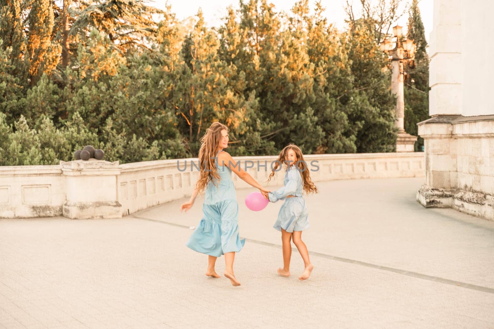 Daughter mother run holding hands. In blue dresses with flowing long hair against the backdrop of a sunset and a white building