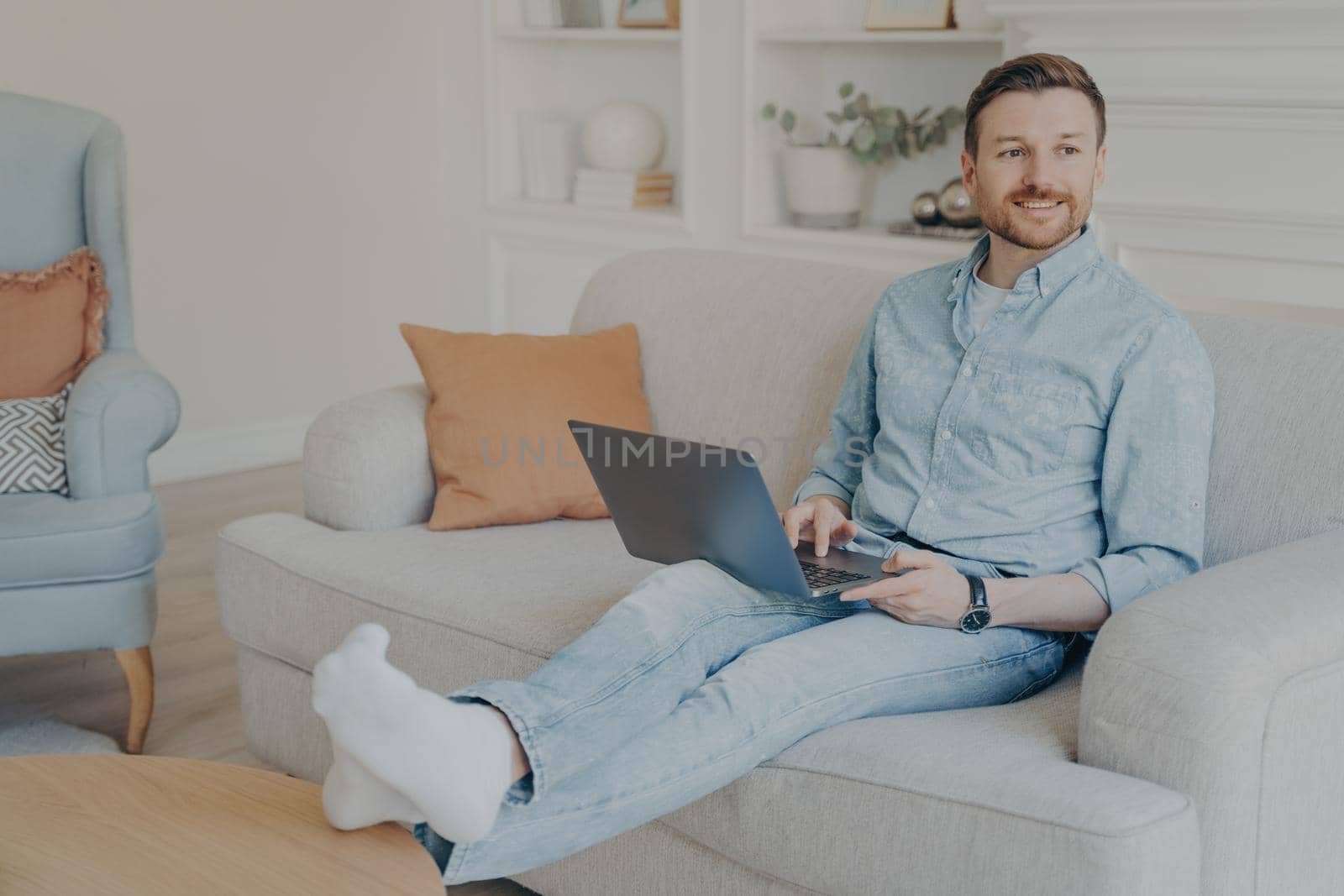 Young man with stubble surfing web on his laptop while sitting on couch, resting legs on coffee table, relaxing after hard work session on project, taking break from working