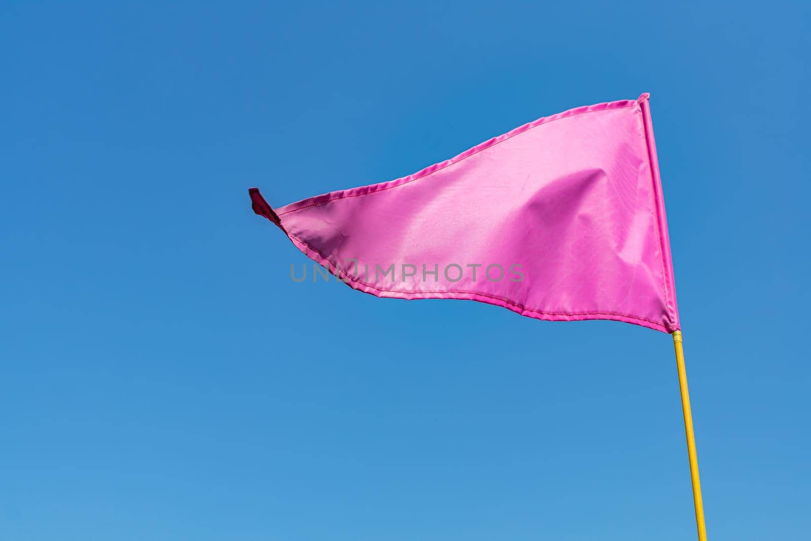 Pink sports flag on a yellow flagpole sways in the wind against a clear blue sky