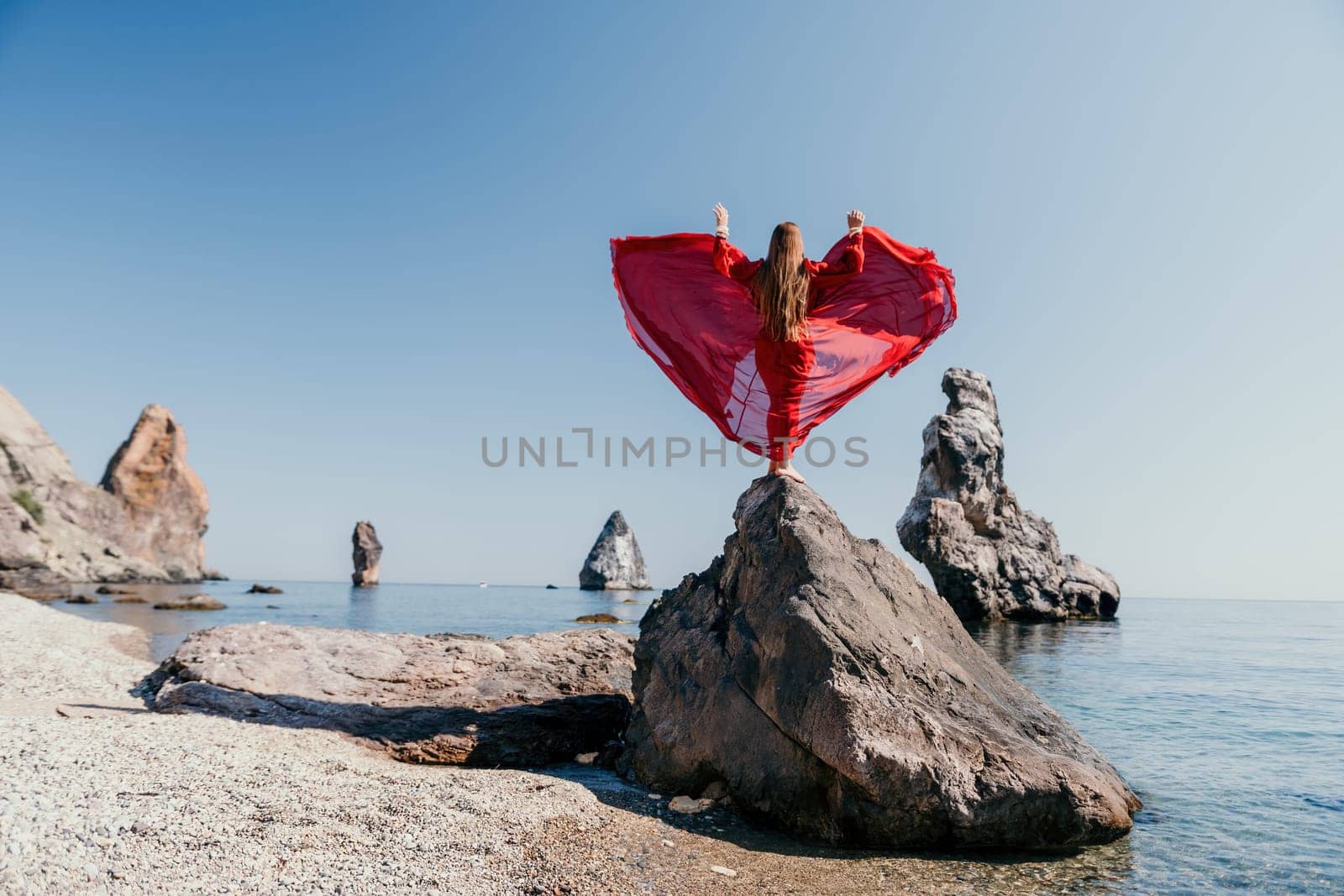 Woman travel sea. Young Happy woman in a long red dress posing on a beach near the sea on background of volcanic rocks, like in Iceland, sharing travel adventure journey by panophotograph