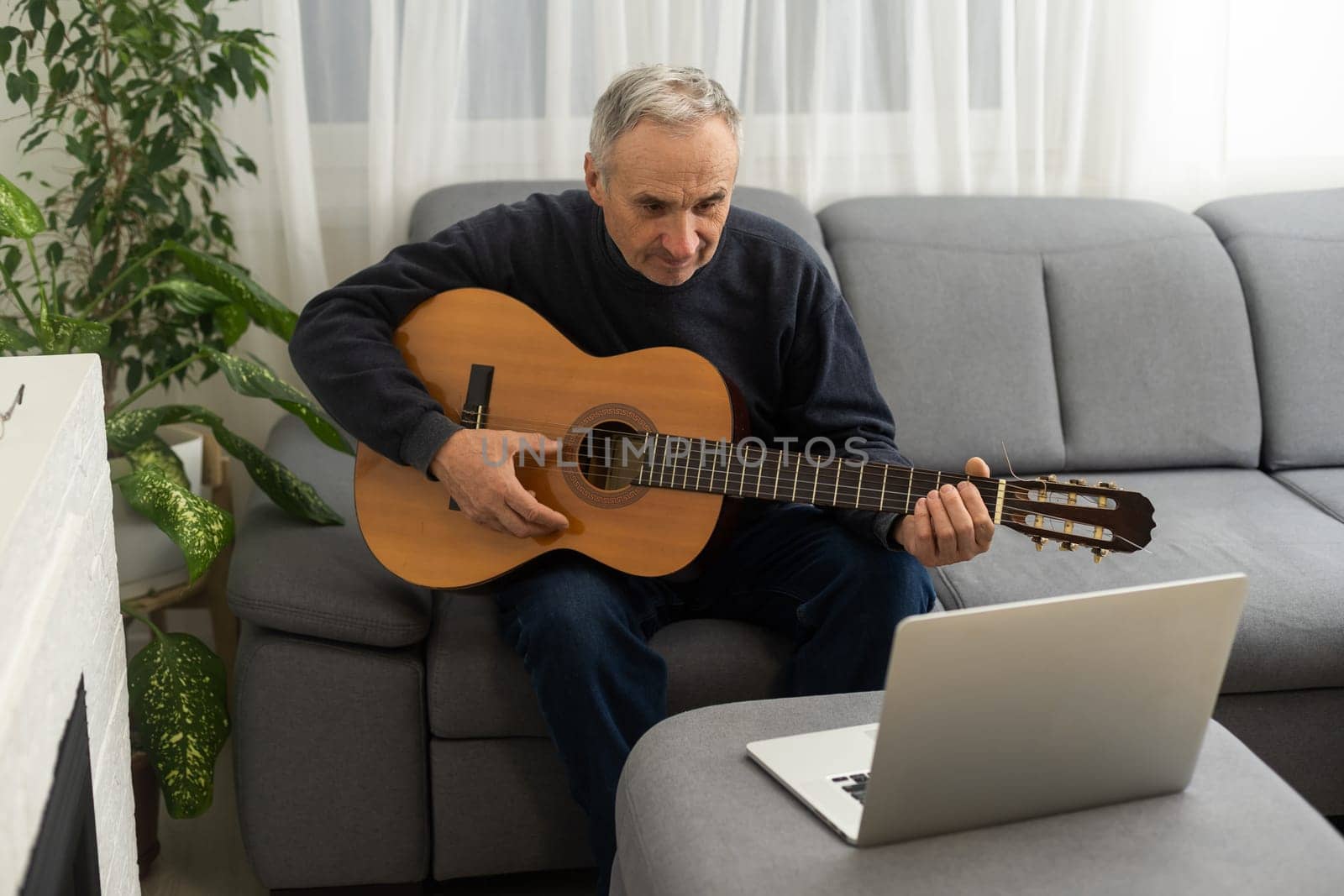 Portrait of senior man in headphones taking online guitar lesson looking at laptop screen. Retired male learning to play guitar watching webinar on computer at home.