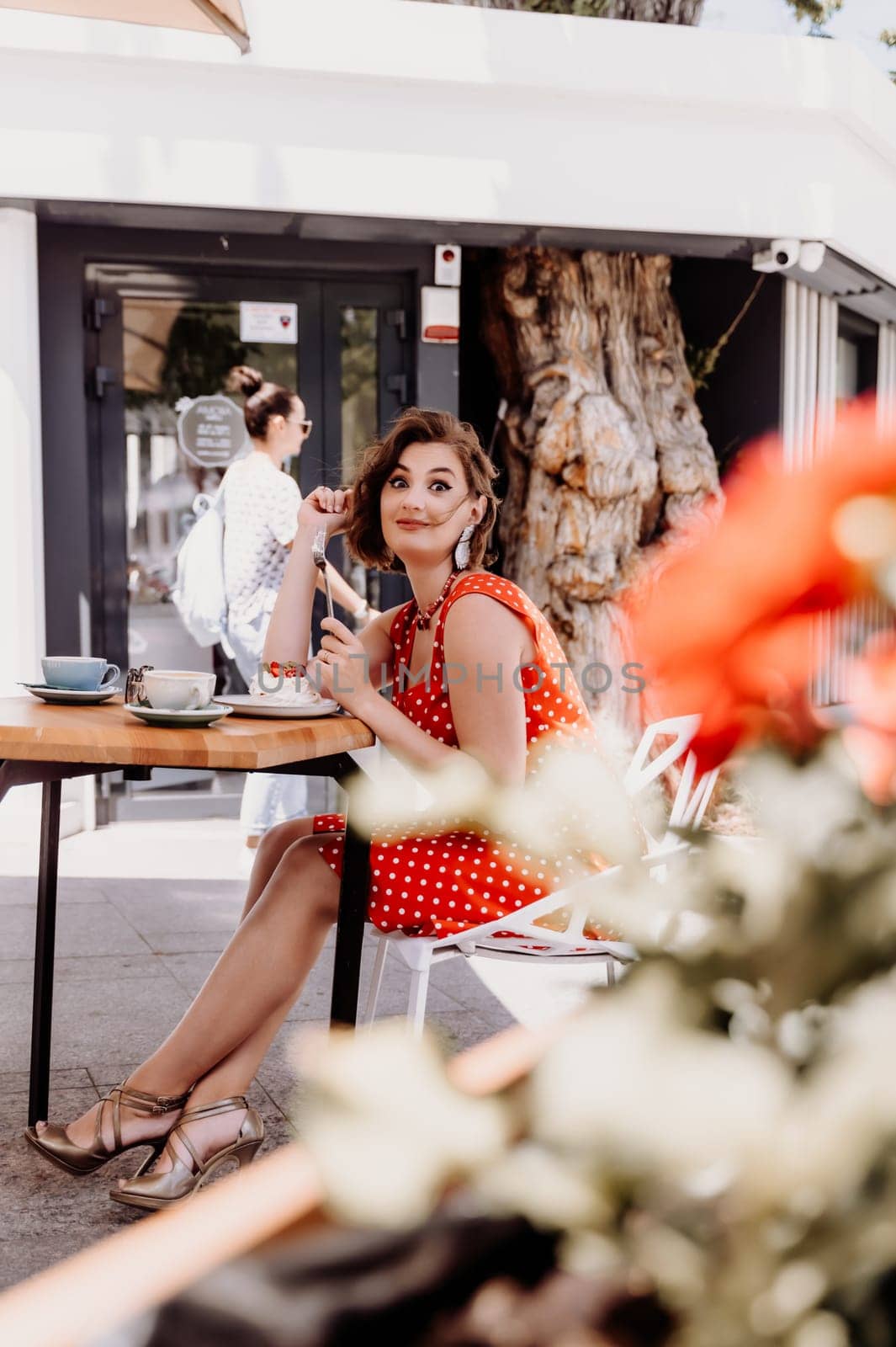 Charming woman in a restaurant, cafe on the street. She sits at the table and eats a cake with a fork. Dressed in a red sundress with white polka dots