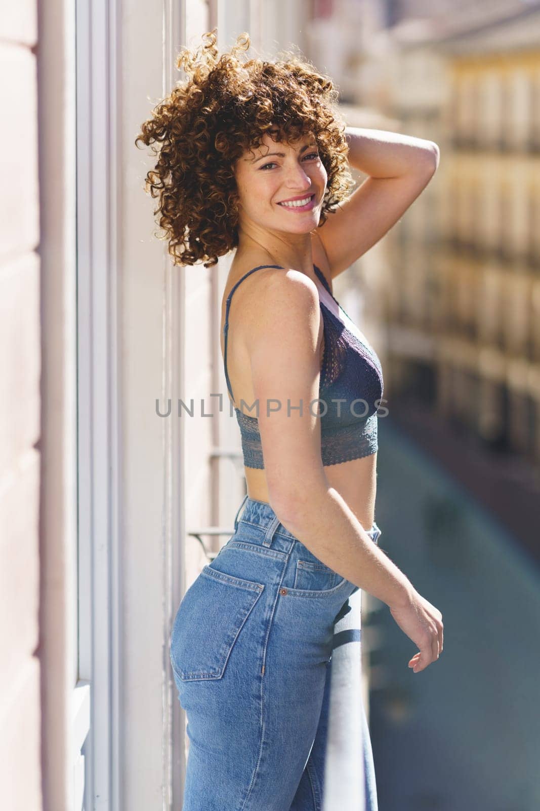 Side view of cheerful young female in bra and jeans smiling while looking at camera and touching curly hair on sunny balcony