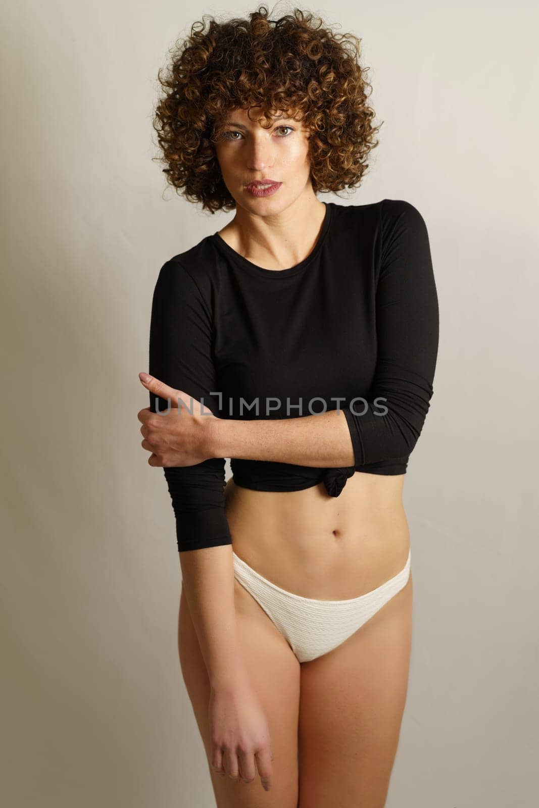 Smiling young slim female model with curly hair,standing with hand folded across chest and touching stretched down arm while looking at camera against gray background