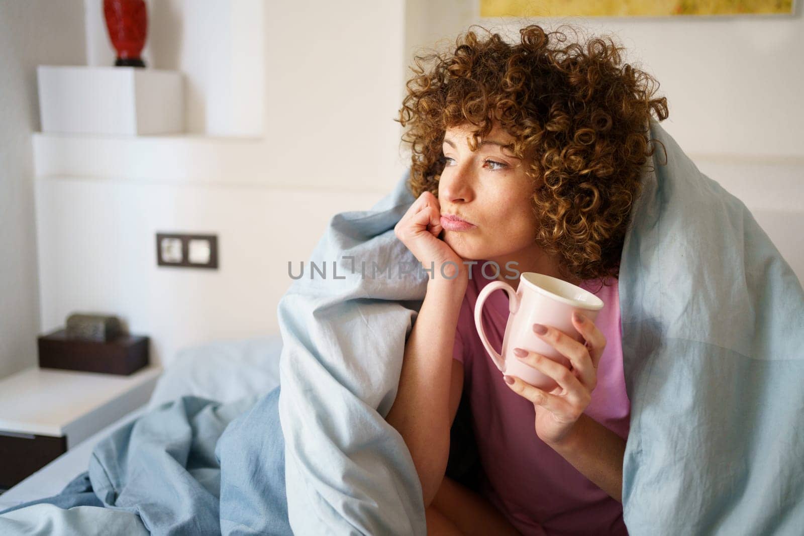 Thoughtful lady looking away and leaning on arm while drinking hot beverage and lounging on bed in morning