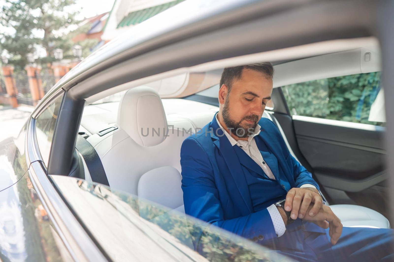 A caucasian man in a blue suit looks at his watch while sitting in the back seat of a car. Business class passenger