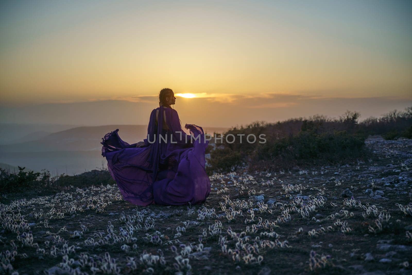 Sunset purple dress woman mountains. Rise of the mystic. sunset over the clouds with a girl in a long purple dress. In the meadow there is a grass dream with purple flowers