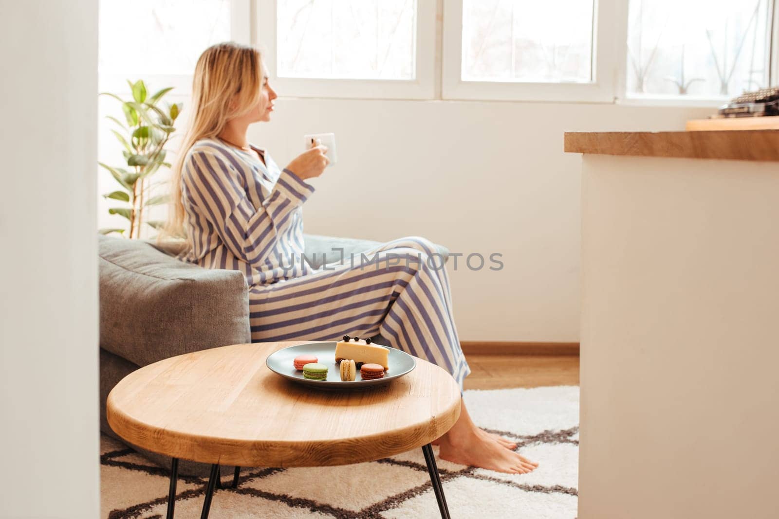 a woman in pajamas in the morning at a table drinks coffee and eats sweets cheesecake sweet macaroni . the concept of breakfast with sweets . side view