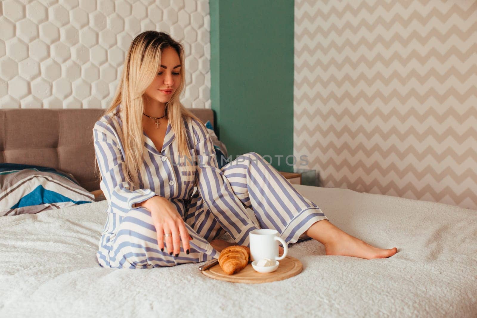 a woman in pajamas drinks coffee with a croissant on the bed . the concept of breakfast in bed.