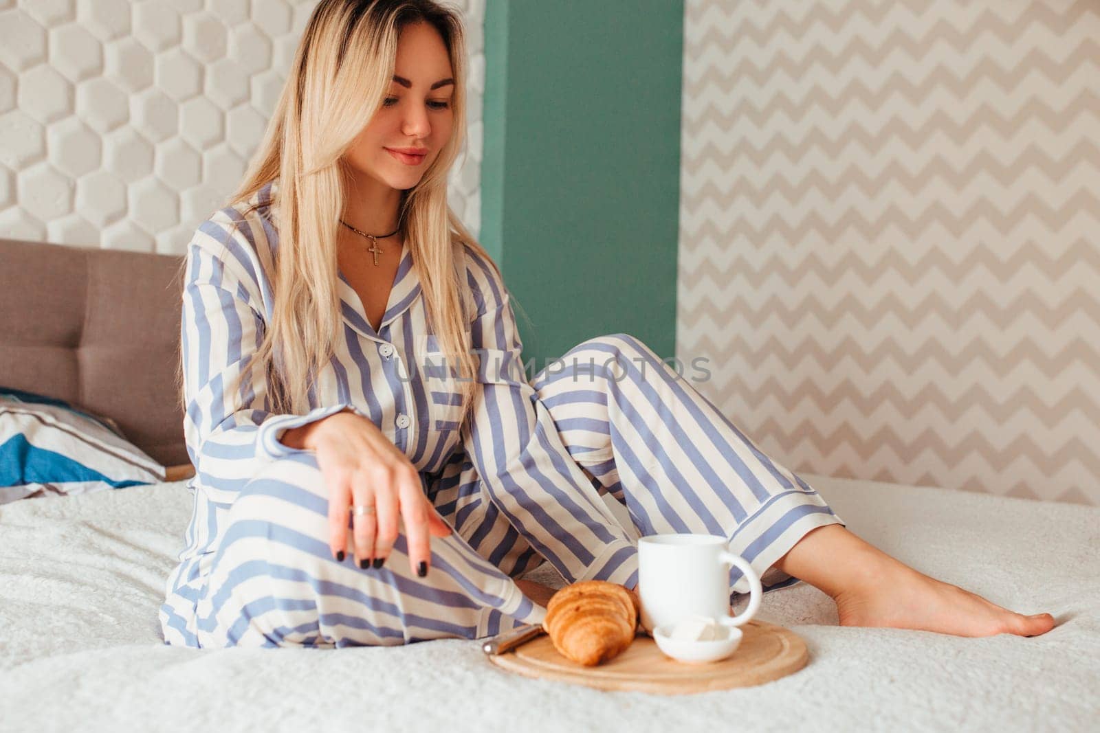 a woman in pyjamas drinks coffee with a croissant on the bed . the concept of breakfast in bed. s