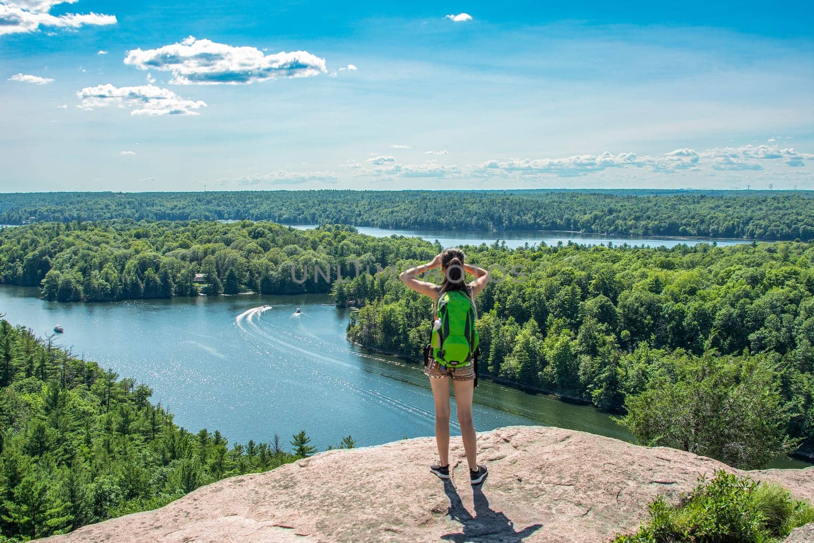 A girl with a backpack stands high on a rock and admires the view of the lakes and forests from above