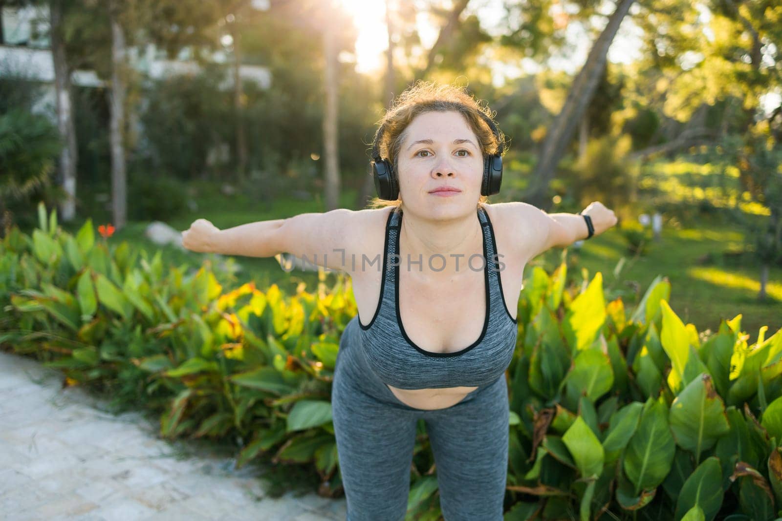 Young pretty smiling plus size woman in sporty top and leggings doing sport and listen to music with headphones in summer morning outdoor by Satura86