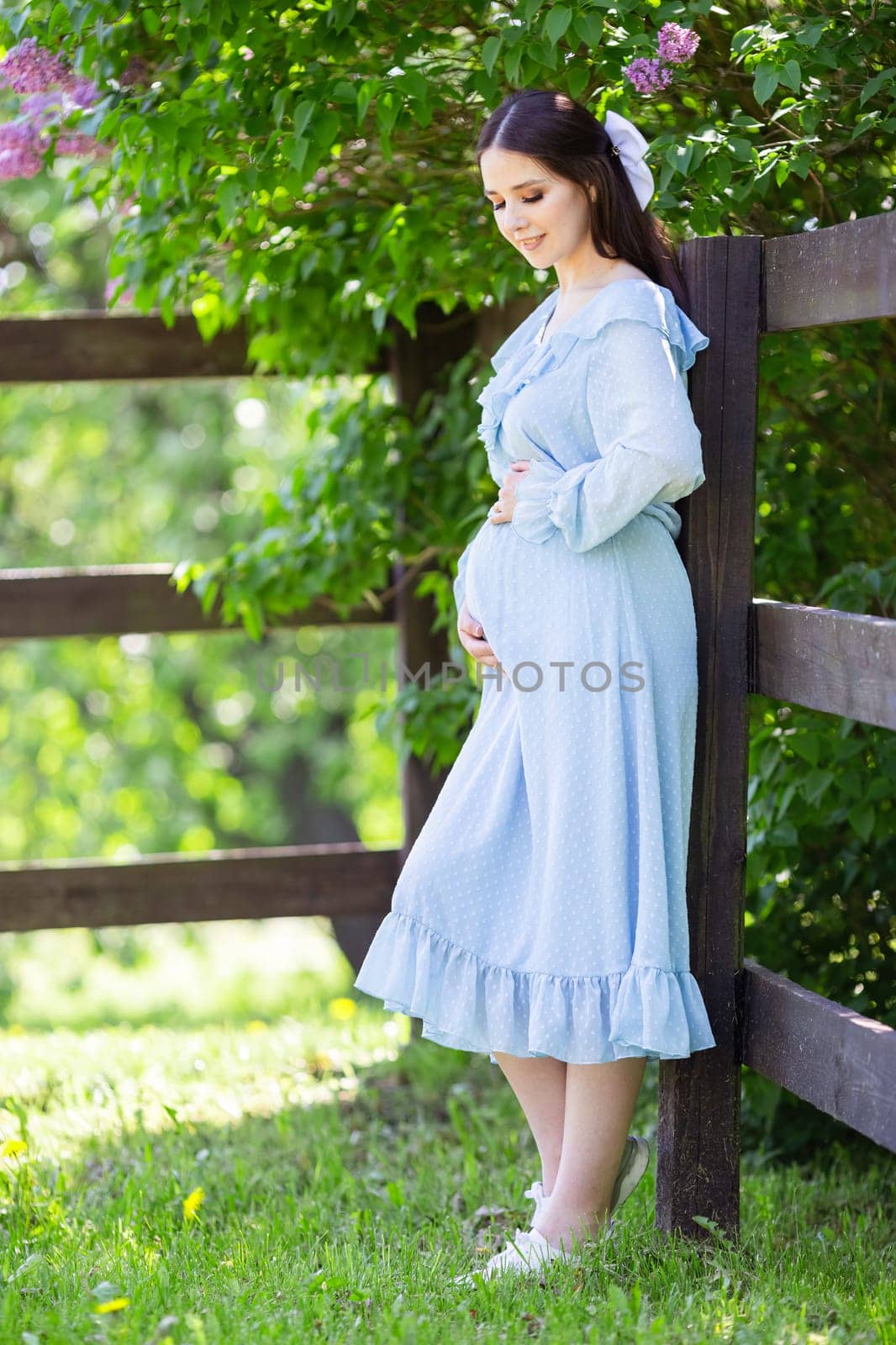Beautiful pregnant young woman with long hair, in a light blue dress stands at the fence, in a lilac garden, on a sunny day. Vertical.Close up. copy space