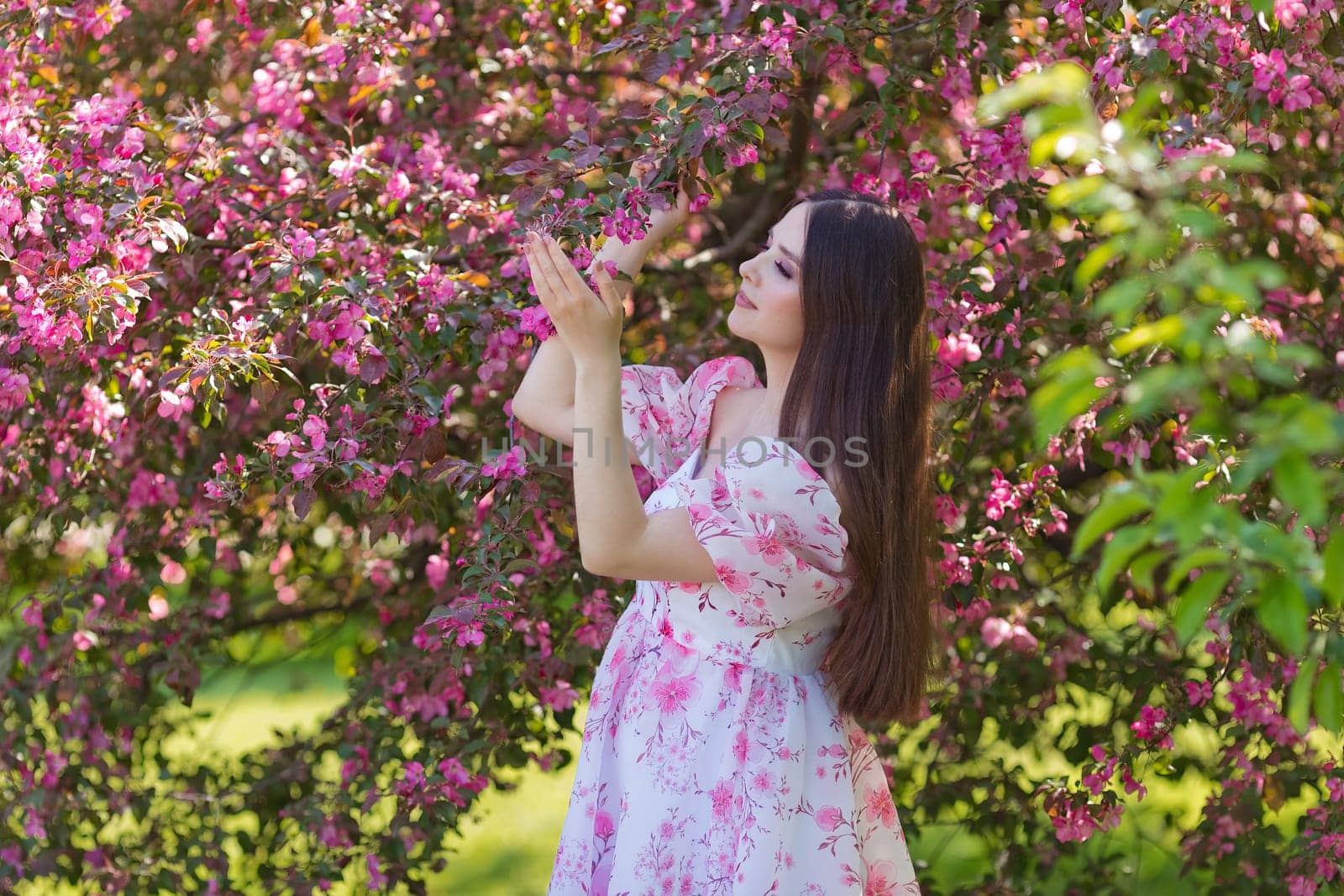 A beautiful girl in a light pink dress standing near pink blooming trees, holding a blooming branch, in the garden. Close up. Copy space