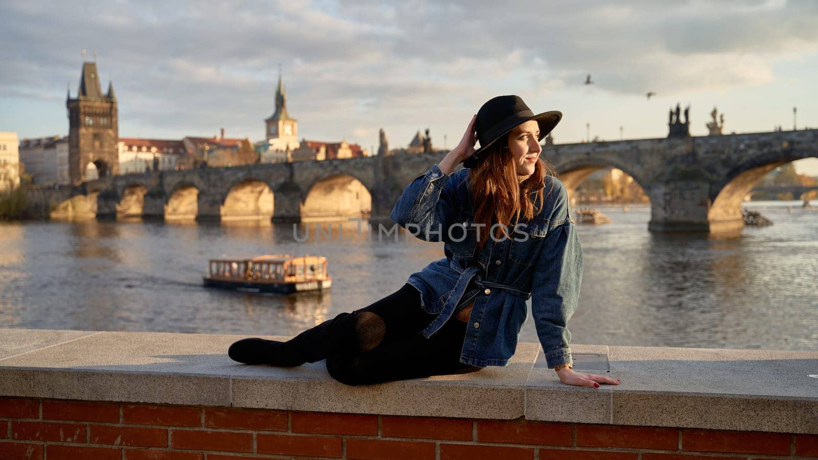 Stylish beautiful young woman wearing black hat sitting on Vltava river shore in Prague with Charles Bridge on background. Elegant retro lady fine art portrait. by berezko