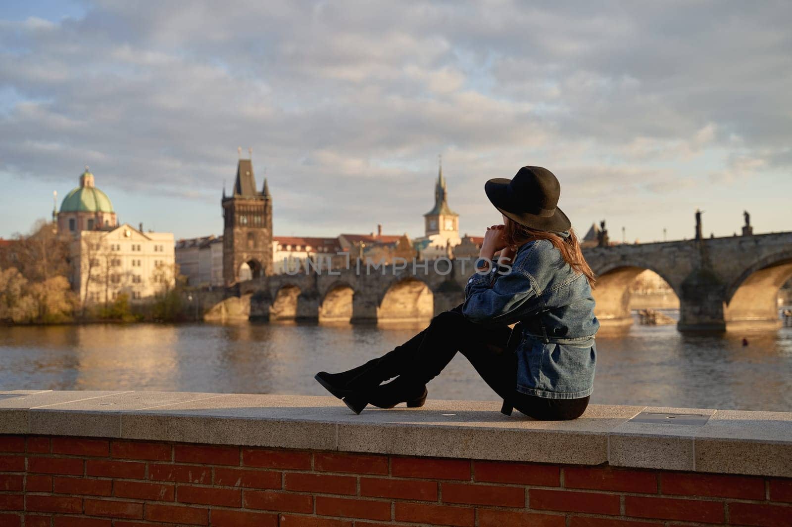 Stylish beautiful young woman wearing black hat sitting on Vltava river shore in Prague with Charles Bridge on background. Elegant retro lady fine art portrait. by berezko