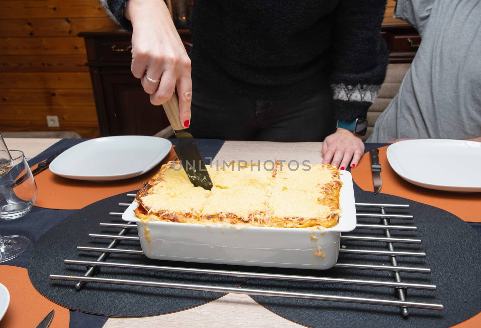 Woman cut with knife hot tasty home baking lasagna in ceramic casserole dish, whole family sit at the table, High quality photo