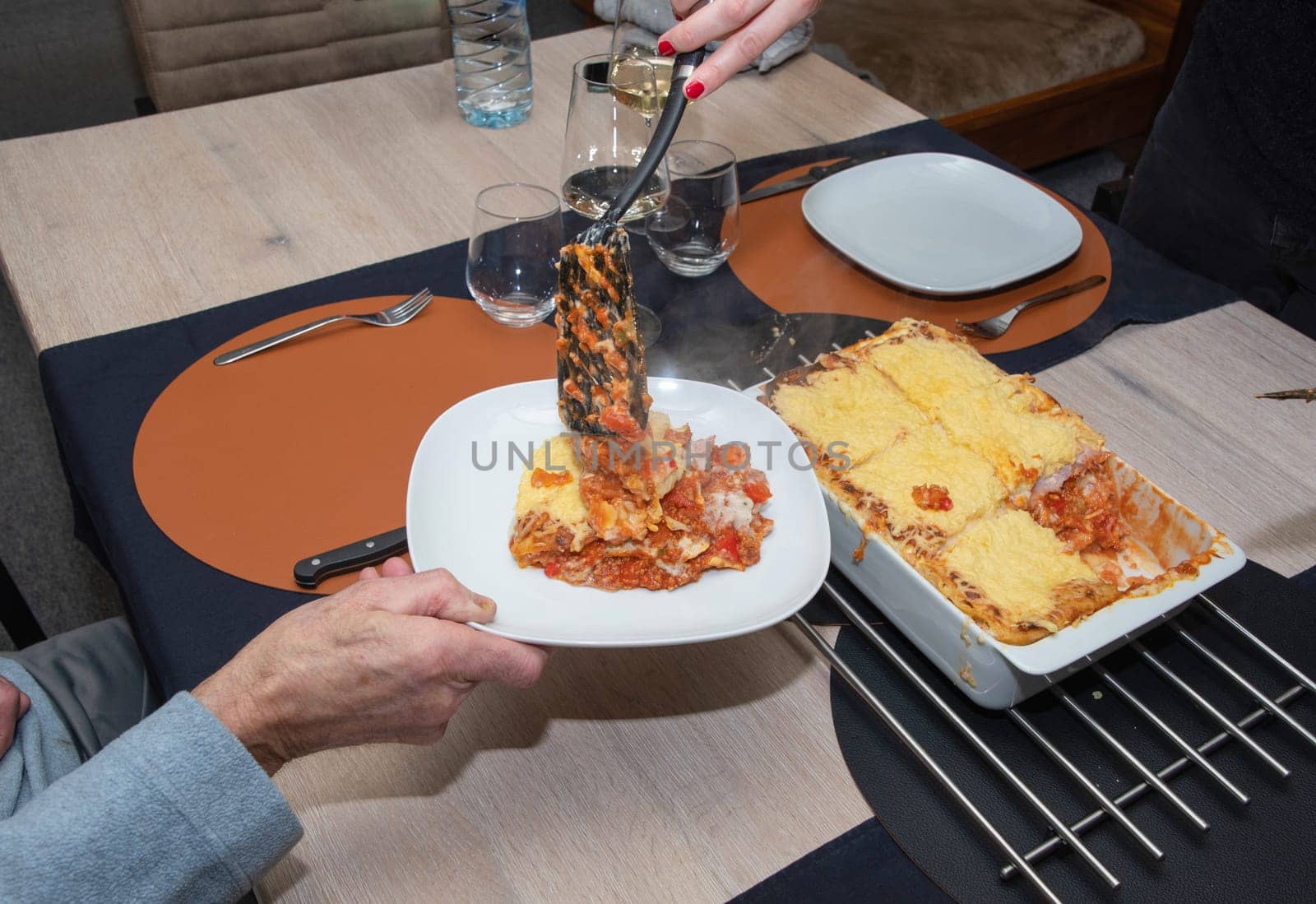 a woman serving hot homemade lasagna on plates to a family member sitting at the table, High quality photo