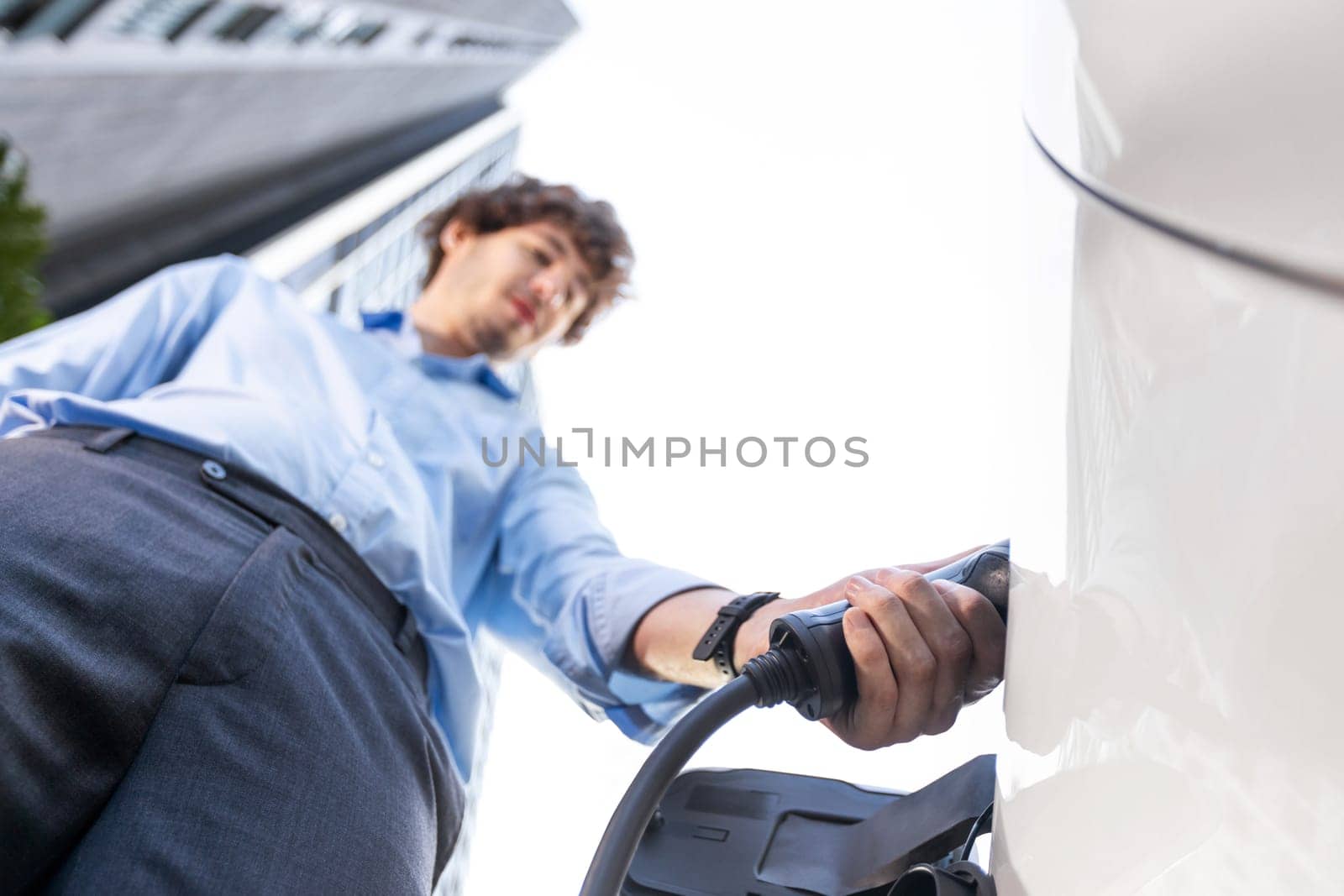 Fisheye view focus on hand insert EV charger plug into electric car with blur background of progressive modern city lifestyle-businessman recharge his EV car with residential building condo background