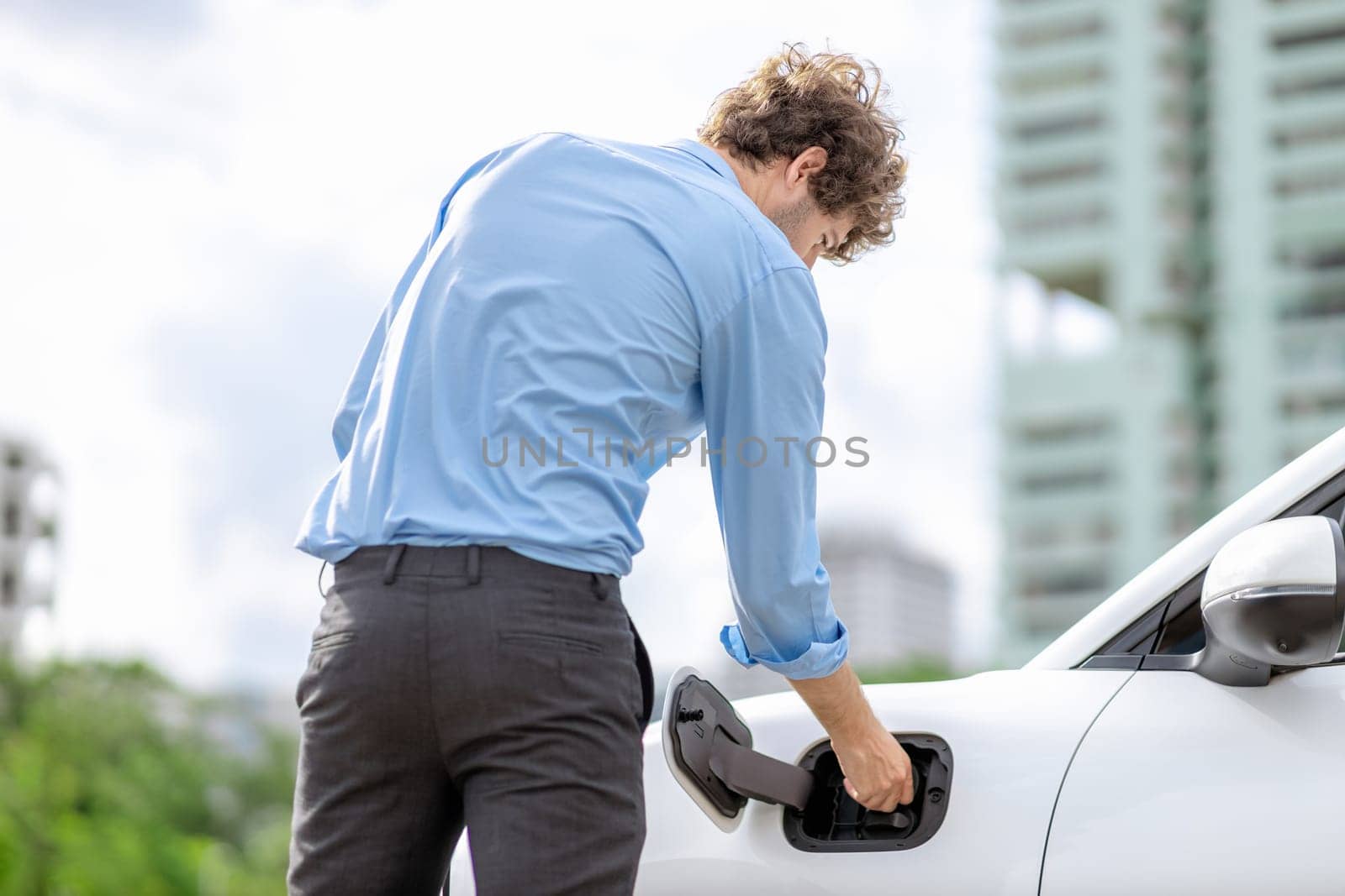 Progressive businessman insert charger plug from charging station to his electric vehicle with apartment condo building in background. Eco friendly rechargeable car powered by sustainable energy.