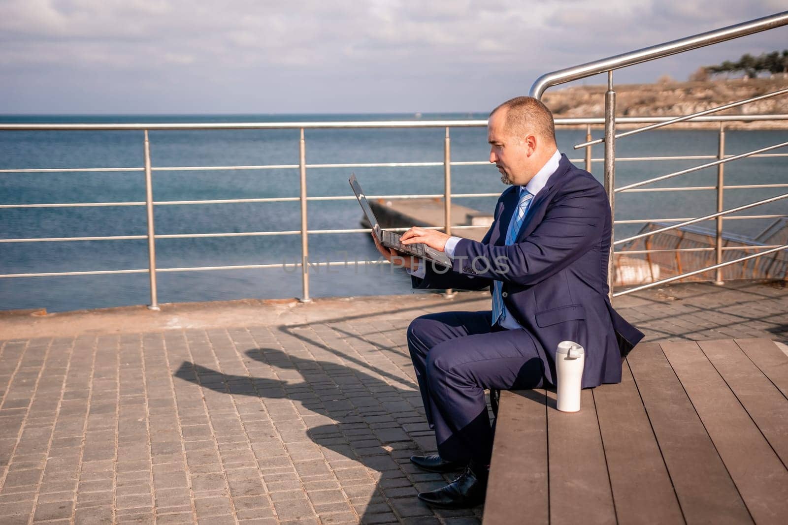 Confident middle age businessman working remotely online, typing on a laptop keyboard while sitting on a beach at sunset. Working remotely on vacation, running an online business from a distance. by panophotograph
