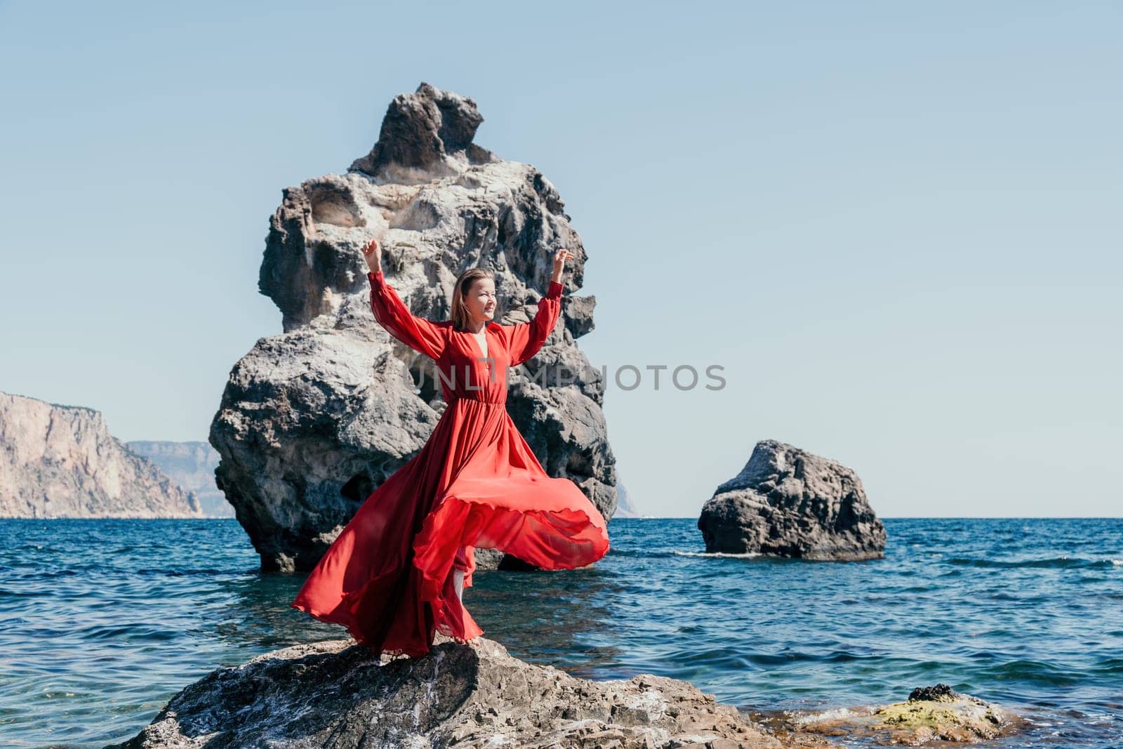 Woman travel sea. Young Happy woman in a long red dress posing on a beach near the sea on background of volcanic rocks, like in Iceland, sharing travel adventure journey